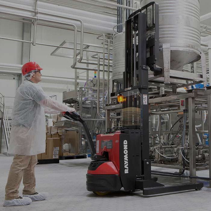 Man in white coat operating a red Raymond Walk-Behind stacker forklift